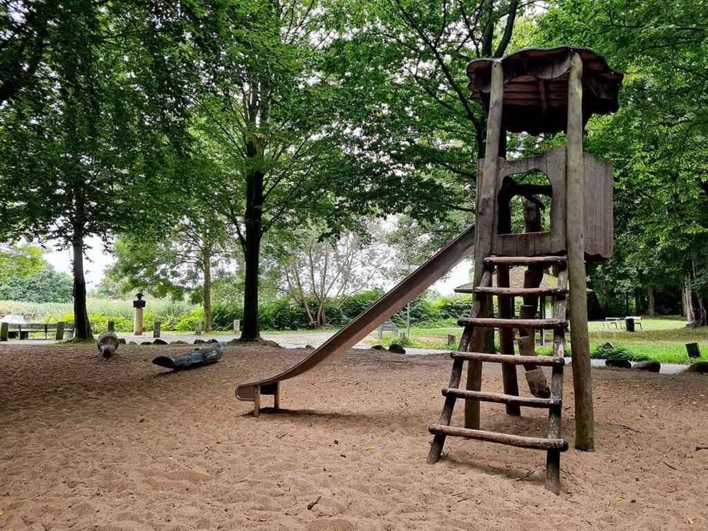 Rutschenturm aus Holz mit Leiter auf einem großen Sandspielbereich im Waldspielplatz von Knoops Park. Links im Hintergrund sind zwei bemalte Holztiere – ein blauer Wal und ein Krokodil – auf dem Sand platziert. Umgeben von hohen, schattenspendenden Bäumen. Im Hintergrund ist ein Weg mit Bänken und der Blick ins Grüne zu erkennen.