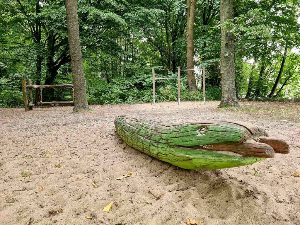 Holzskulptur in Form eines grünen Krokodils auf dem Sandspielplatz im Knoops Park. Im Hintergrund stehen Turnstangen und ein Balancierbalken aus Holz zwischen Bäumen. Die gesamte Szene ist von dichtem Wald umgeben und vermittelt eine natürliche, ruhige Atmosphäre.