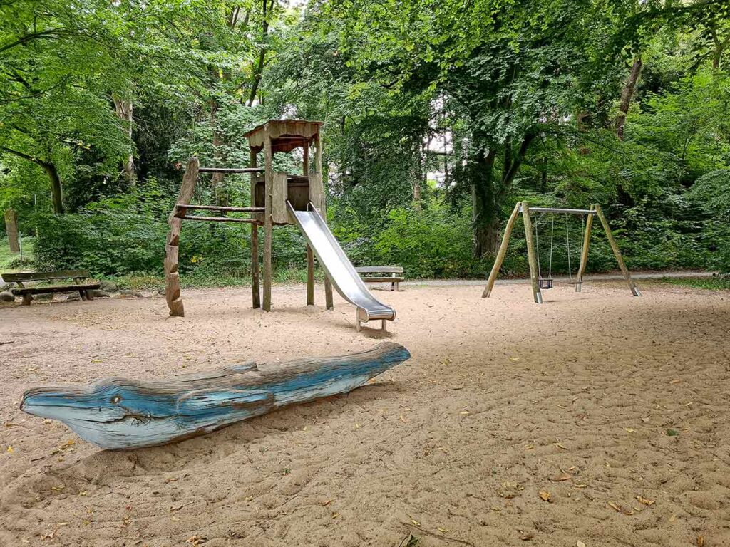 Blick auf den Spielplatz im Knoops Park mit einer großen Rutsche, einer Holzskulptur in Form eines blauen Delfins im Vordergrund und einer Doppelschaukel im Hintergrund. Der gesamte Spielplatz liegt auf sandigem Boden und ist von hohen, dicht bewachsenen Bäumen umgeben.