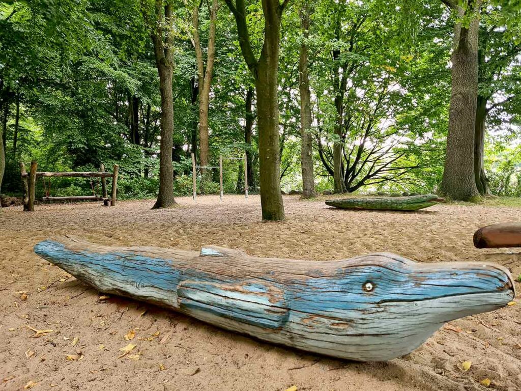 uf dem Spielplatz im Knoops Park liegt ein geschnitzter, blau bemalter Wal aus Holz im Sandbereich. Im Hintergrund sind weitere Holzspielgeräte sowie ein Kletterbereich zwischen Bäumen zu sehen. Der gesamte Spielplatz liegt idyllisch unter hohen, schattenspendenden Bäumen.