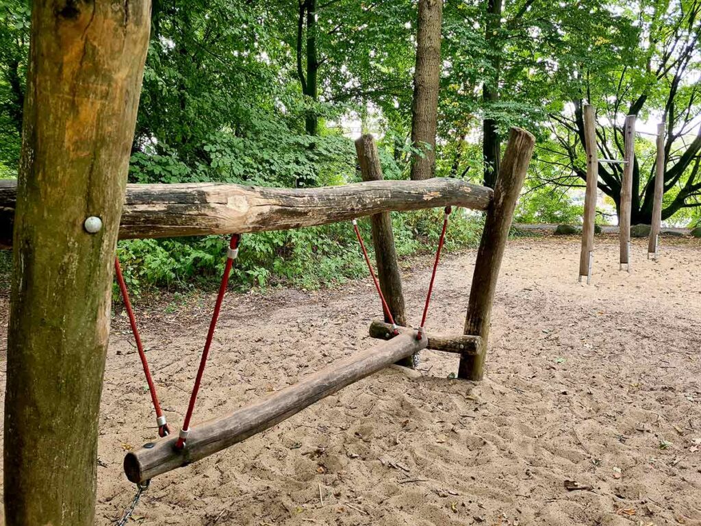 Ein Balanciergerät aus naturbelassenen Holzstämmen auf dem Spielplatz im Knoops Park. Es besteht aus einem schwebenden, an Seilen aufgehängten Balken, der von zwei Holzrahmen gehalten wird. Der Boden ist mit Sand bedeckt, im Hintergrund stehen weitere Kletterelemente zwischen hohen Bäumen.
