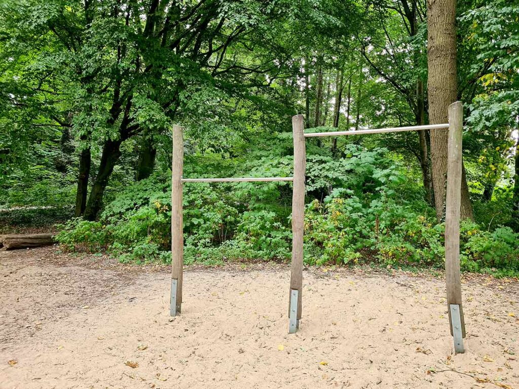 Zwei Turnstangen in unterschiedlicher Höhe auf dem Spielplatz im Knoops Park. Die Metallstangen sind zwischen naturbelassenen Holzpfosten montiert. Der Boden ist mit Sand bedeckt, dahinter dichtes, grünes Gebüsch und hohe Bäume.