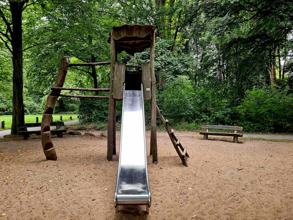 Eine hohe Edelstahlrutsche an einem rustikalen Kletterturm aus naturbelassenem Holz. Links eine gebogene Kletterwand, rechts eine schräge Holzleiter. Der Boden ist sandig, im Hintergrund stehen Bänke und dichter Baumbestand – typische Parkatmosphäre.