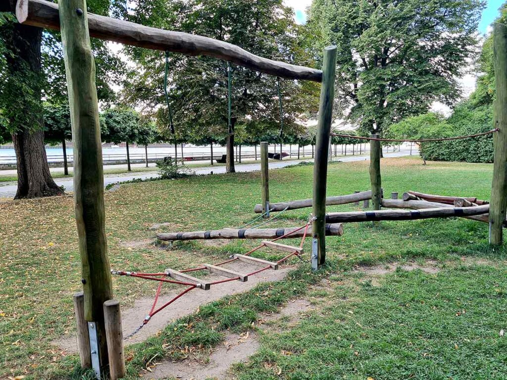 Teil des Kletter- und Balancierbereichs im Stadtgarten Vegesack. Zu sehen ist eine Hängebrücke aus roten Seilen und Holzsprossen, eingebettet in eine grüne Wiesenfläche. Im Hintergrund verlaufen Spazierwege und dahinter die Uferpromenade an der Weser, gesäumt von Bäumen.