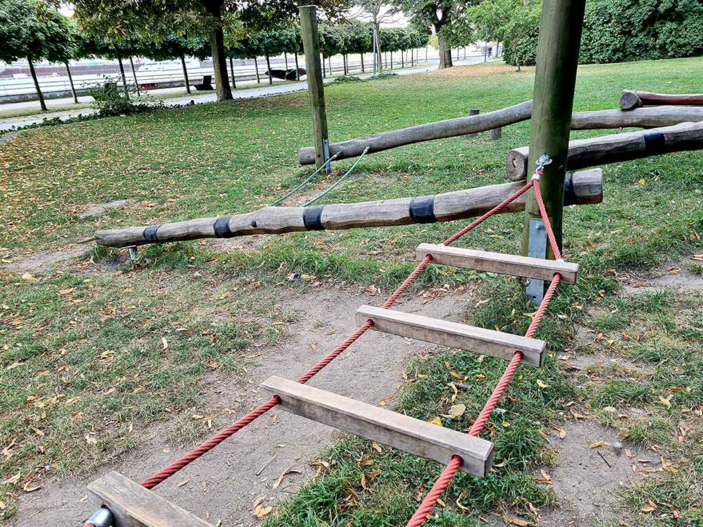 Nahaufnahme eines Kletter- und Balancierelements im Stadtgarten Vegesack. Im Vordergrund eine Hängebrücke aus roten Seilen und Holzsprossen, im Hintergrund geschwungene Balancierbalken aus Holz. Die Spielgeräte stehen auf einer Rasenfläche nahe der Weserpromenade.