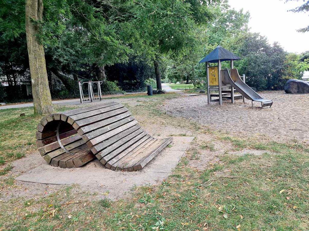 Übersicht über den Spielplatz im Stadtgarten Vegesack mit einer kleinen Rutsche mit Podest und einem geschwungenen Klettertunnel aus Holz im Vordergrund. Im Hintergrund sind Fitnessgeräte, ein Baum und dichter Bewuchs zu erkennen. Der Boden ist teilweise mit Sand und Rasen bedeckt.