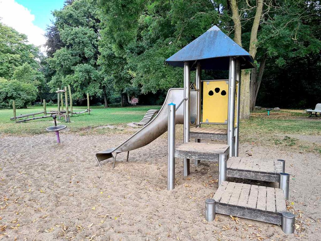 Spielplatz im Stadtgarten Vegesack mit einem kleinen Spielturm aus Metall und Holz, einer Rutsche und Podesttreppen. Im Hintergrund sind Balancierbalken, ein Drehspielgerät und grüne Rasenflächen zu sehen. Der Bereich ist von großen Bäumen umgeben, die für viel Schatten sorgen.