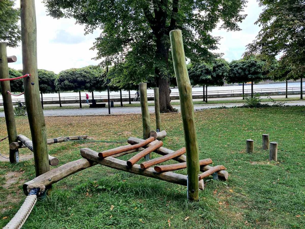 Kletterelement aus Holz auf dem Spielplatz im Stadtgarten Vegesack mit Blick auf die Weser im Hintergrund. Die Konstruktion besteht aus schräg angeordneten Rundhölzern und ist Teil eines größeren Balancier- und Kletterbereichs auf einer Rasenfläche. Im Hintergrund sind ein Weg, Bäume mit dichtem Laub und das Wasser zu sehen.