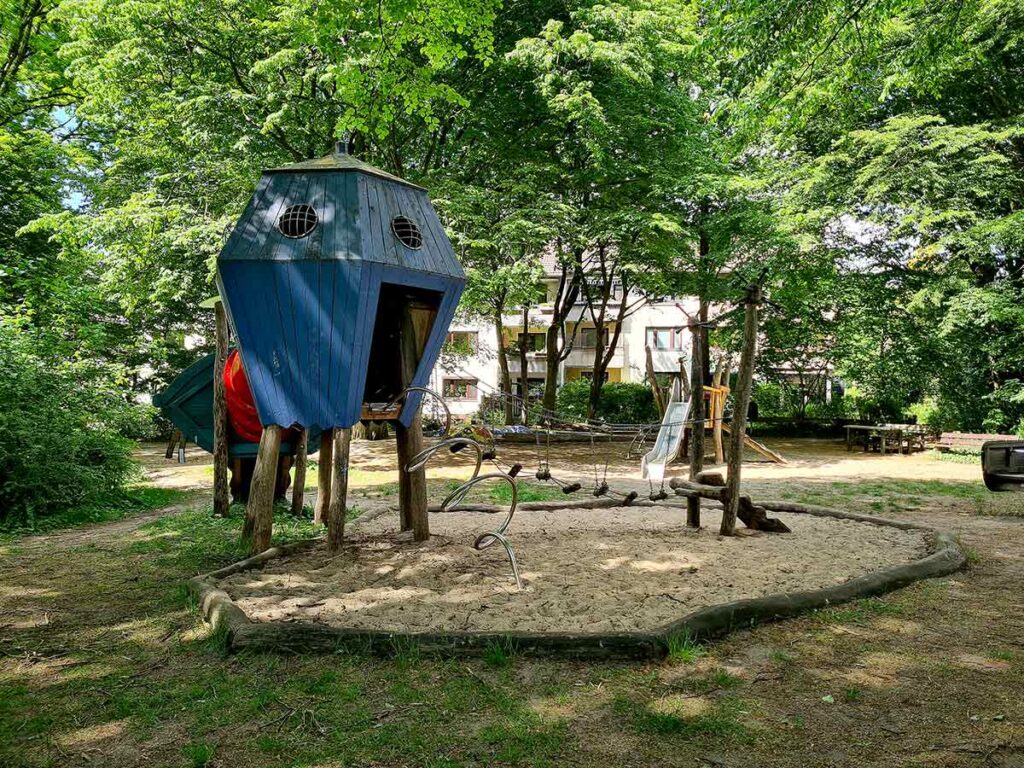 Spielplatz im Grünen mit markanter, blauer Spielhütte auf Stelzen, Kletterelementen aus Holz und Seilen sowie Rutsche im Hintergrund – umgeben von großen Bäumen und Wohnhäusern.