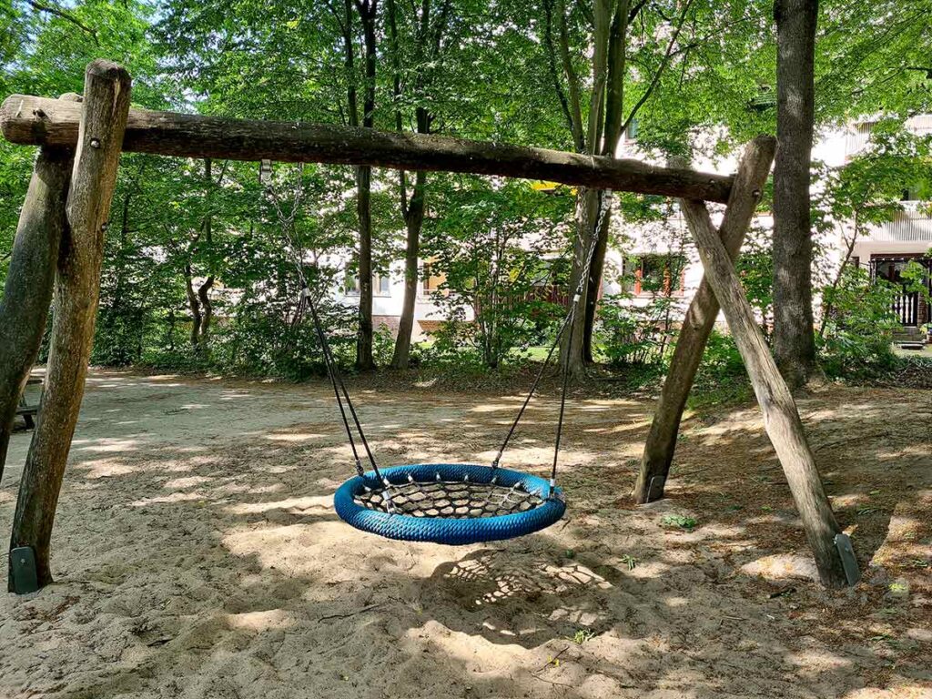 Nestschaukel mit blauem Rand unter schattigen Bäumen, befestigt an einem robusten Holzgestell auf sandigem Boden, im Hintergrund Wohnhäuser.