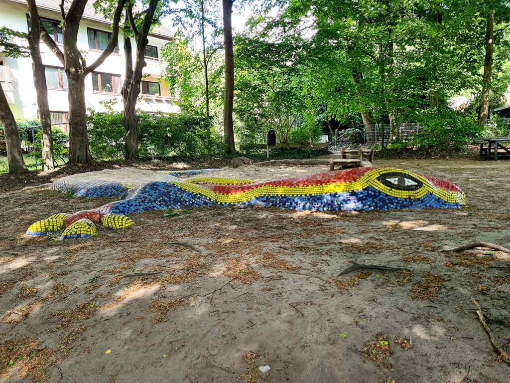 Bunte Mosaik-Eidechse aus Keramikkacheln in Rot, Gelb, Blau und Schwarz, halb in den Boden eingelassen, als fantasievolles Spielelement im schattigen Bereich des Spielplatzes zwischen Bäumen.
