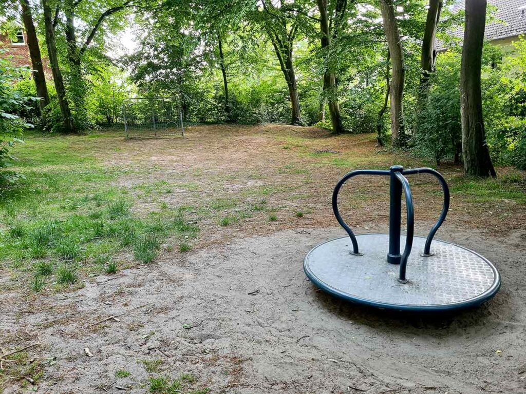 Metallkarussell auf sandigem Untergrund am Rand einer Lichtung, im Hintergrund ein kleines Fußballtor und dichter Baumbestand – naturnaher Bereich des Spielplatzes.