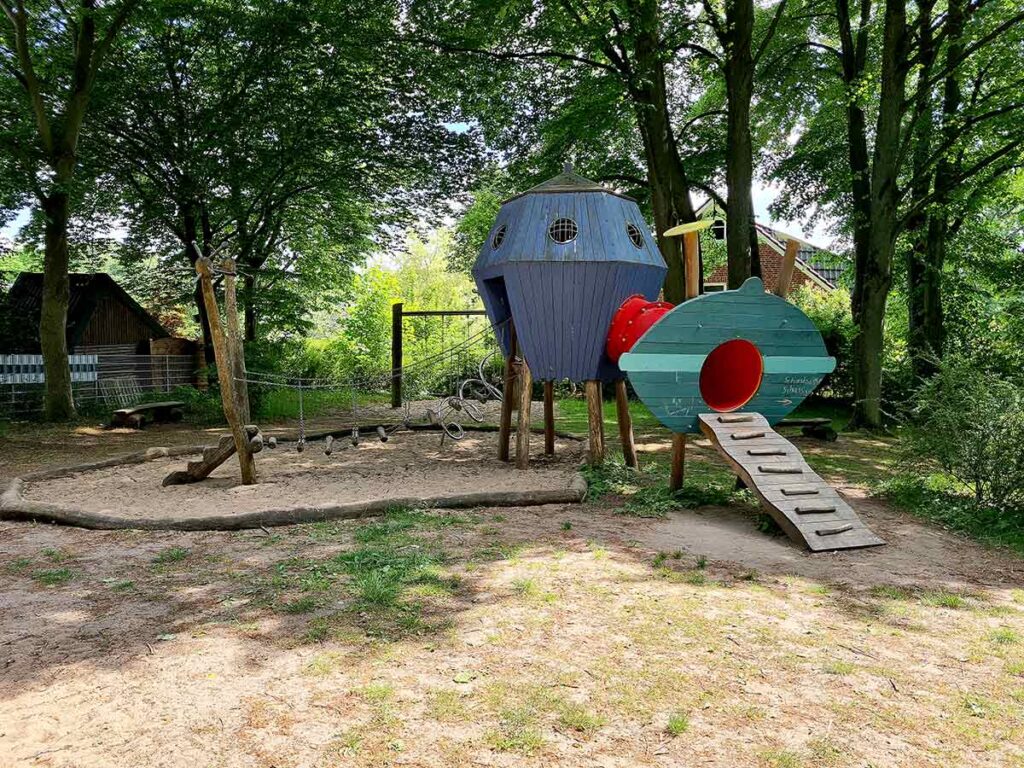 Fantasievolles Klettergerüst in Form eines blauen Ufos mit rotem Tunnel und grünem Einstiegselement, daneben ein Balancier- und Kletterbereich mit Seilen, alles eingebettet in eine schattige Spielfläche unter hohen Bäumen.