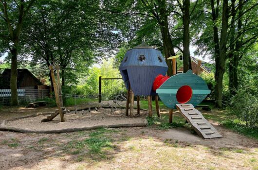 Fantasievolles Klettergerüst in Form eines blauen Ufos mit rotem Tunnel und grünem Einstiegselement, daneben ein Balancier- und Kletterbereich mit Seilen, alles eingebettet in eine schattige Spielfläche unter hohen Bäumen.