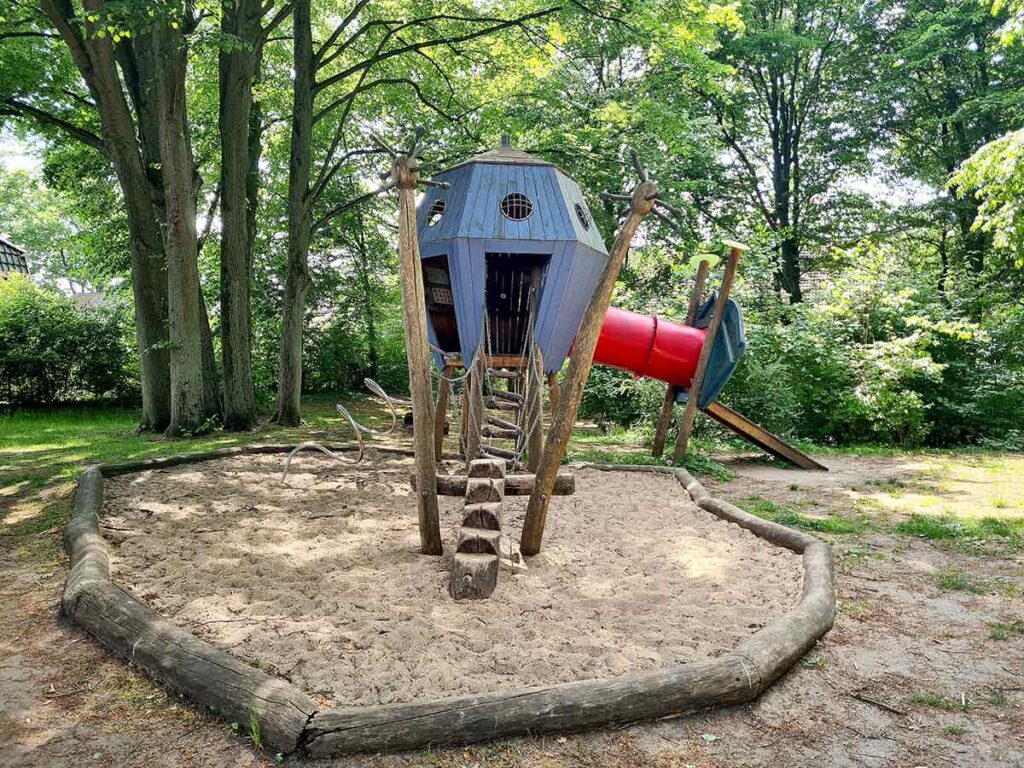 Kletterbereich mit einem blauen Ufo-ähnlichen Spielhaus, das über eine rote Tunnelrutsche verfügt. Der Zugang erfolgt über Kletterelemente im Sandbereich, umgeben von schattenspendenden Bäumen.
