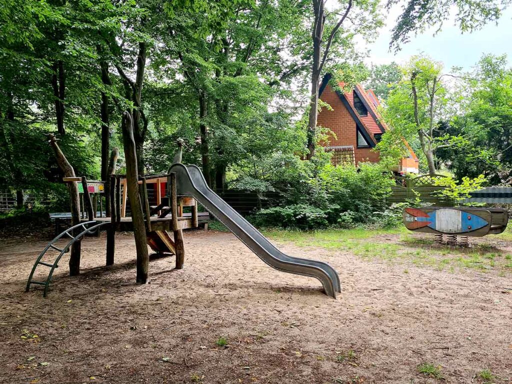Naturbelassenes Klettergerüst mit Rutsche, Kletternetz und Holzplattform auf einem Sanduntergrund. Rechts im Bild eine Wippe in Fischform, im Hintergrund ein markantes Haus mit rotem Satteldach. Der Spielbereich ist von Bäumen umgeben.