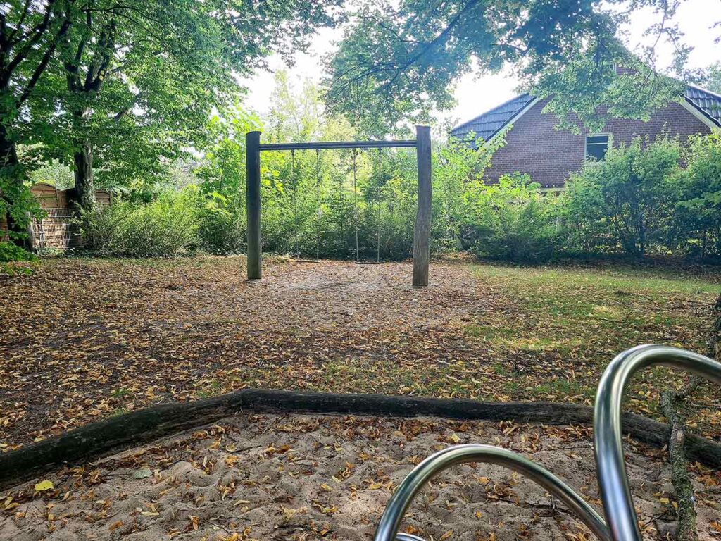 Schlichte Holzschaukel mit zwei Sitzen auf sandigem Boden in einem von Bäumen und Büschen umgebenen Bereich. Im Vordergrund ein Ausschnitt eines Spielkarussells aus Metall. Im Hintergrund ein rotes Backsteinhaus mit dunklem Walmdach.