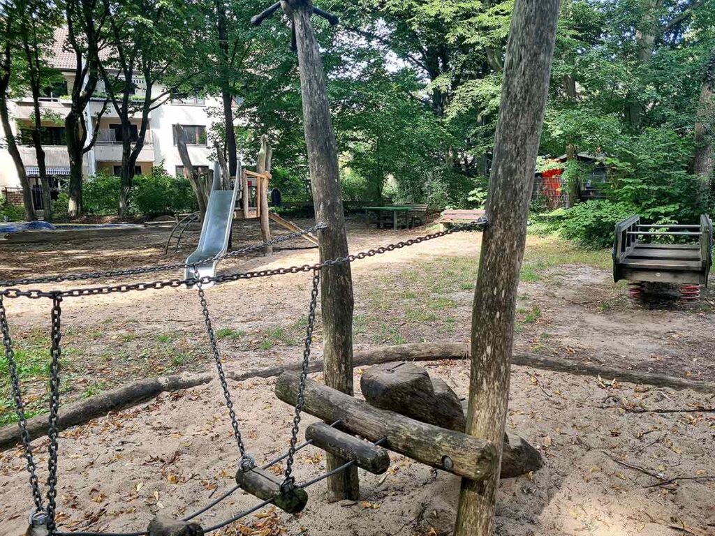Balancierbrücke aus Ketten und Holzstämmen in einem naturbelassenen Sandbereich. Im Hintergrund eine Rutsche, Kletterelemente und ein Spielhäuschen. Rechts eine kleine Federwippe in Form eines Holzwagens. Umgeben von Bäumen und Wohnhäusern.