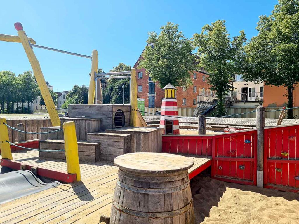 Heckbereich des maritimen Spielschiffs in Vegesack mit Holzpodesten, Leuchtturm-Attrappe, Kletterelementen und einem Fass im Sand.