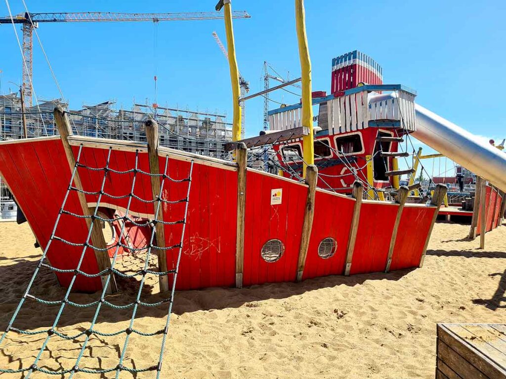 Seitenansicht des großen roten Spielschiffs in Vegesack mit Kletternetz, Bullaugen und Edelstahlrutsche, im Hintergrund Baugerüste und Kräne
