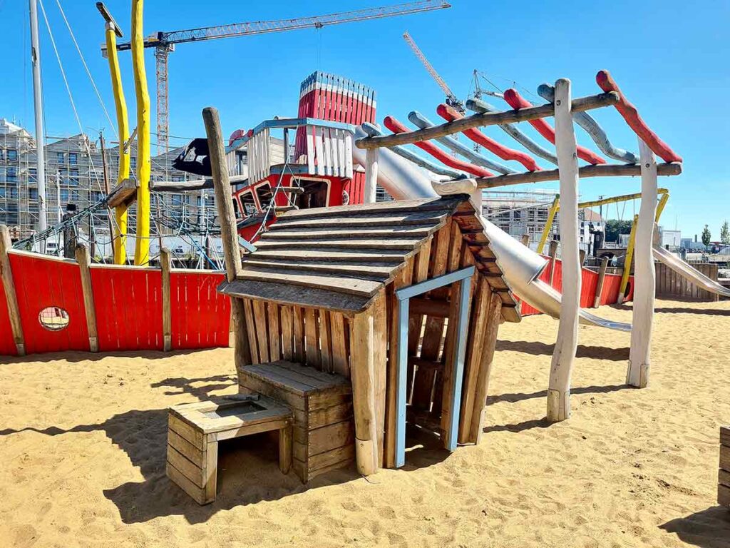 Kleines Holzspielhaus mit Bank im Sand, im Hintergrund das große Spielschiff mit Rutschen, Takelage und buntem Kletterbogen.