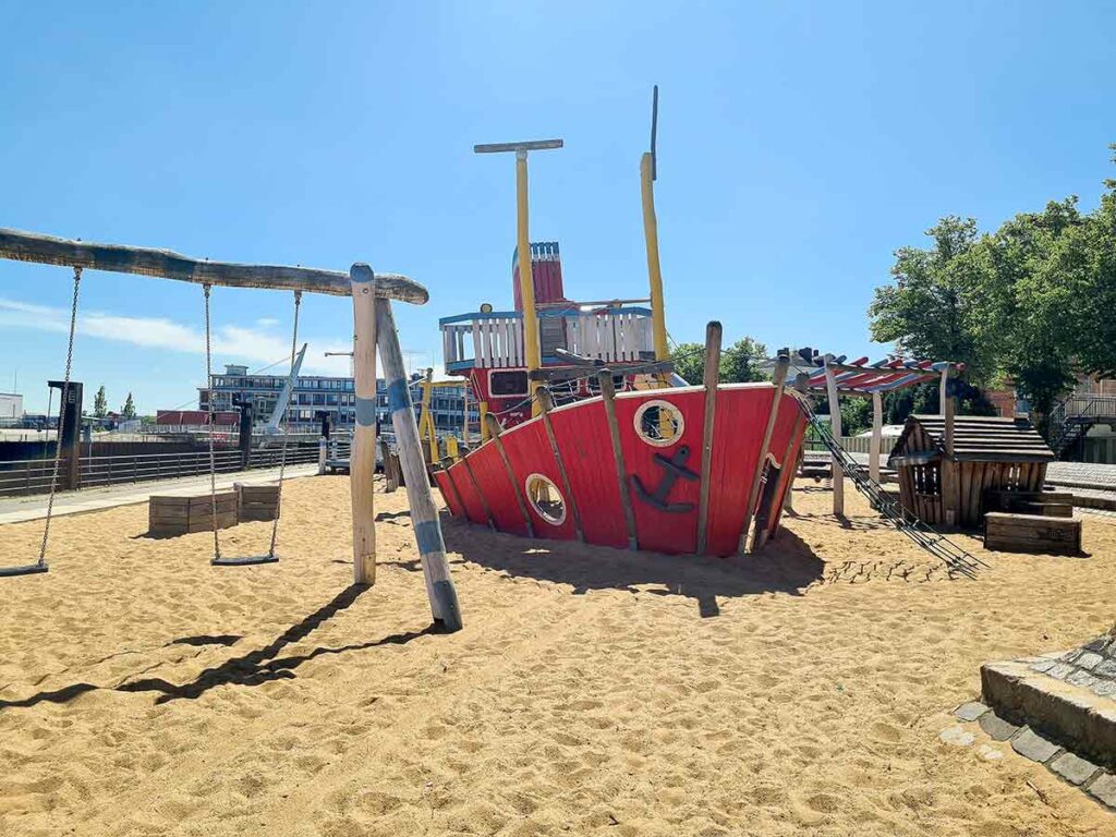 Gesamtansicht des Spielbereichs an der Vegesacker Promenade mit Schaukeln, großem Spielschiff im Sand und kleinem Holzhaus, im Hintergrund moderne Wohngebäude.