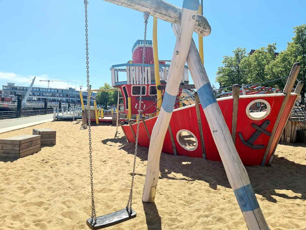 Vorderansicht einer Holzschaukel auf dem sandigen Boden des Spielplatzes, im Hintergrund das rote Spielschiff mit Bullaugen und Anker, direkt an der Promenade in Vegesack.