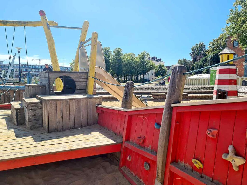 Detailaufnahme eines Spielbereichs mit Holzelementen, kleiner Rutsche, Kletterwand mit bunten Griffen und Leuchtturm-Attrappe auf dem maritimen Spielplatz in Vegesack.