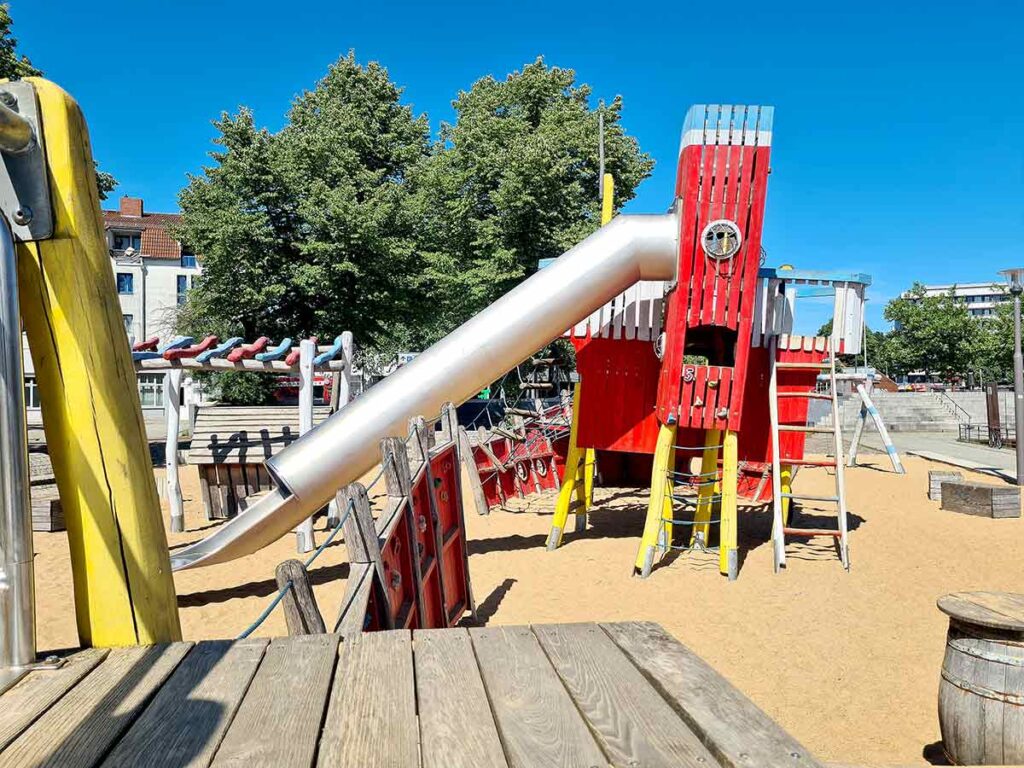 Blick auf das große rote Spielschiff mit Edelstahlrutsche, Kletternetzen und Leitern auf dem Sandspielplatz an der Weserpromenade in Vegesack.
