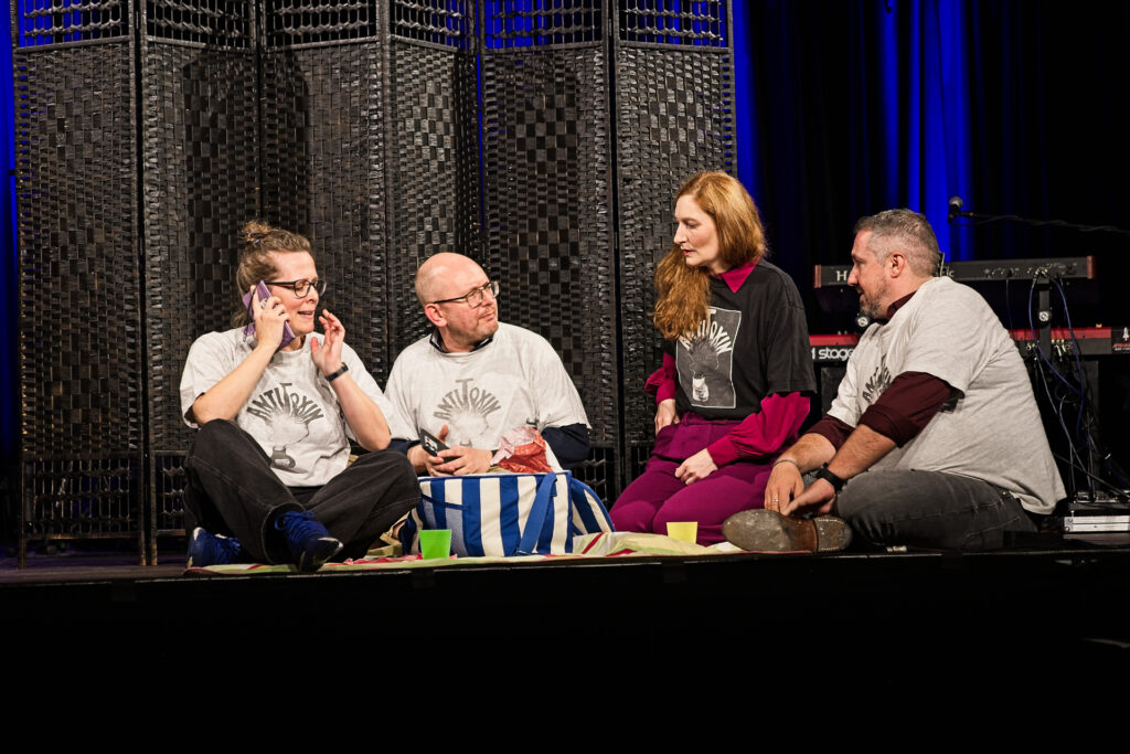 Vier Erwachsene sitzen auf einer Bühne im Kreis, teils mit AntiToxin-T-Shirts, vor einem schwarz geflochtenen Bühnenhintergrund. Sie spielen eine Szene nach: eine Familie geht in den Park, alle haben Handys.
