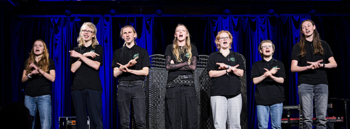 Sieben Jugendliche des aktuellen AntiToxin-Ensembles stehen auf einer Bühne in einer Reihe und singen. Alle tragen schwarze Oberteile mit Logo-Aufdruck. Ihre Arme sind in einer synchronen Geste überkreuzt. Im Hintergrund sind Bühnenvorhang, Instrumente und Lichttechnik zu sehen. Das Foto entstand während der Jubiläumsgala 2025.