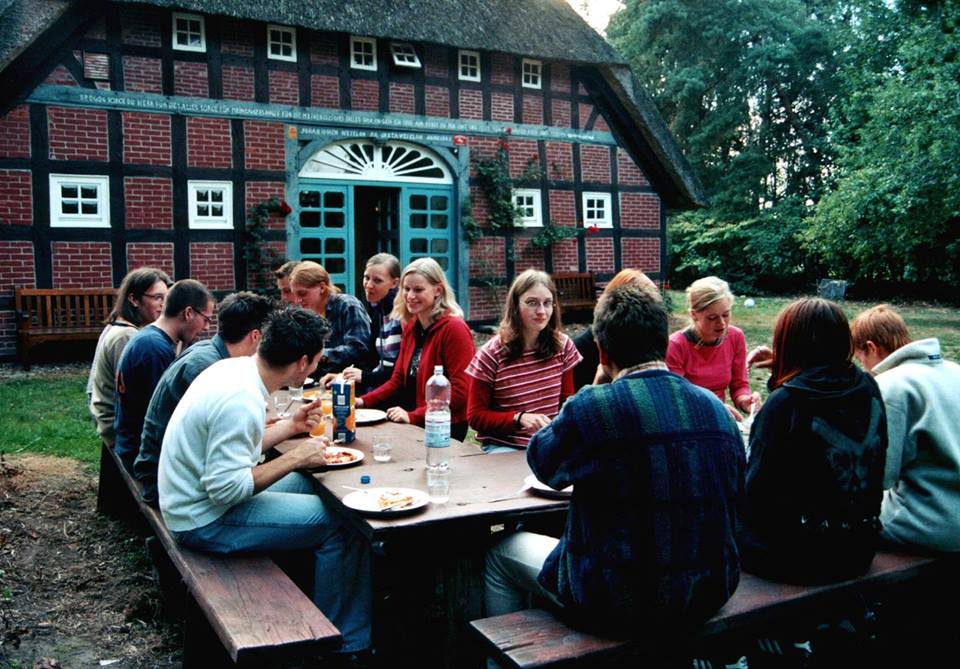 Gruppe junger Menschen beim gemeinsamen Essen an einem Holztisch im Garten vor einem historischen Fachwerkhaus mit Reetdach in Borchel. Das Foto zeigt Mitglieder des Schülerkabaretts AntiToxin im Jahr 2002 während einer Arbeitsphase zur Programmentwicklung.
