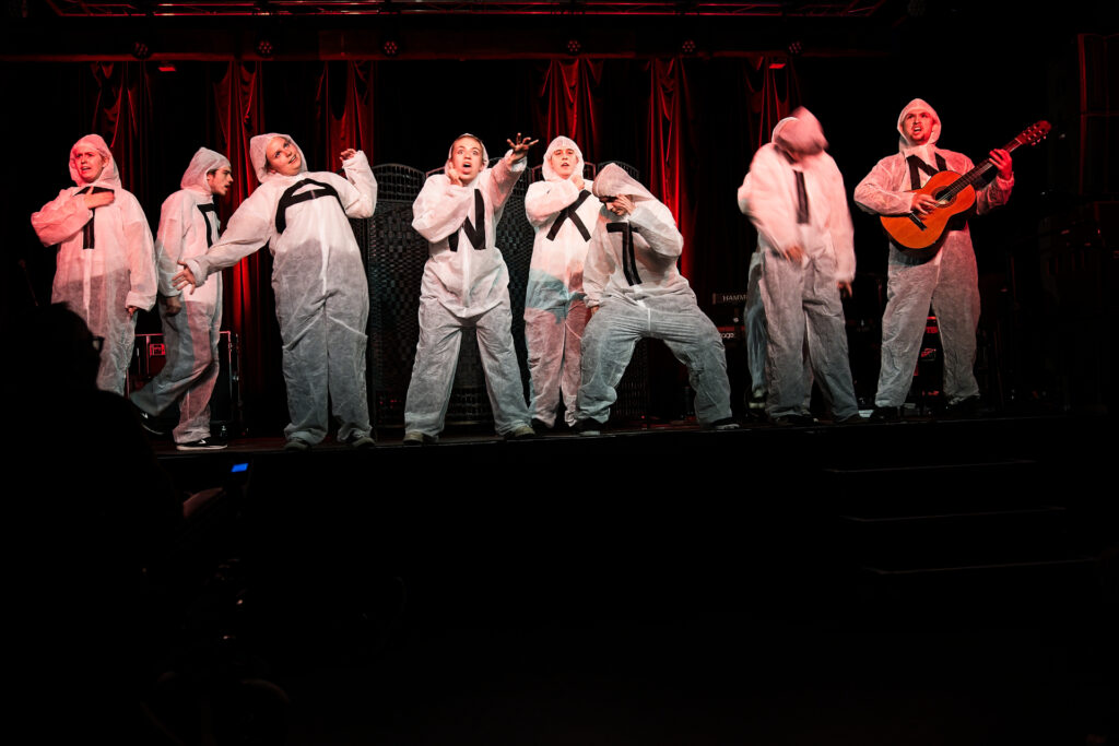 Szene aus dem AntiToxin-Jubiläumsprogramm 2023: Acht Darsteller*innen in weißen Schutzanzügen mit schwarzen Kreuzen performen auf einer rot beleuchteten Bühne eine parodistische Gesangseinlage. Eine Person spielt Gitarre.