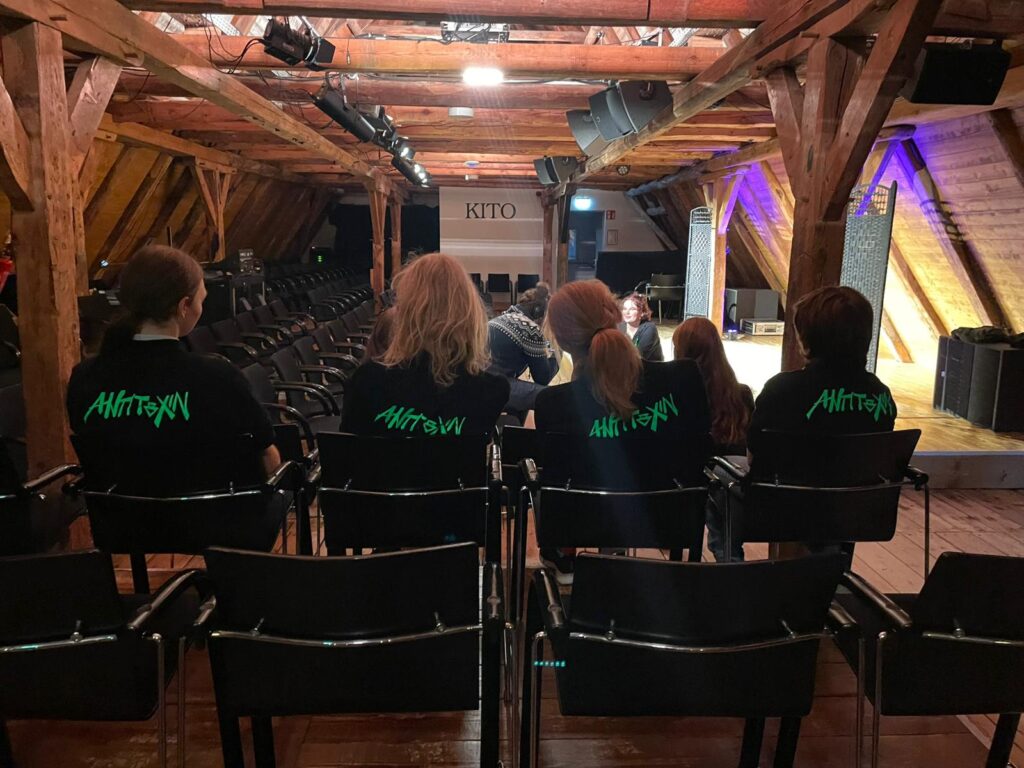 Aufnahme von Hinten: mehrere Jugendliche sitzen im KITO auf den Bänken und blicken zur Bühne, auf den Rücken ihrer T-Shirts ist der giftgrüne Schriftzug "AntiToxin" zu lesen