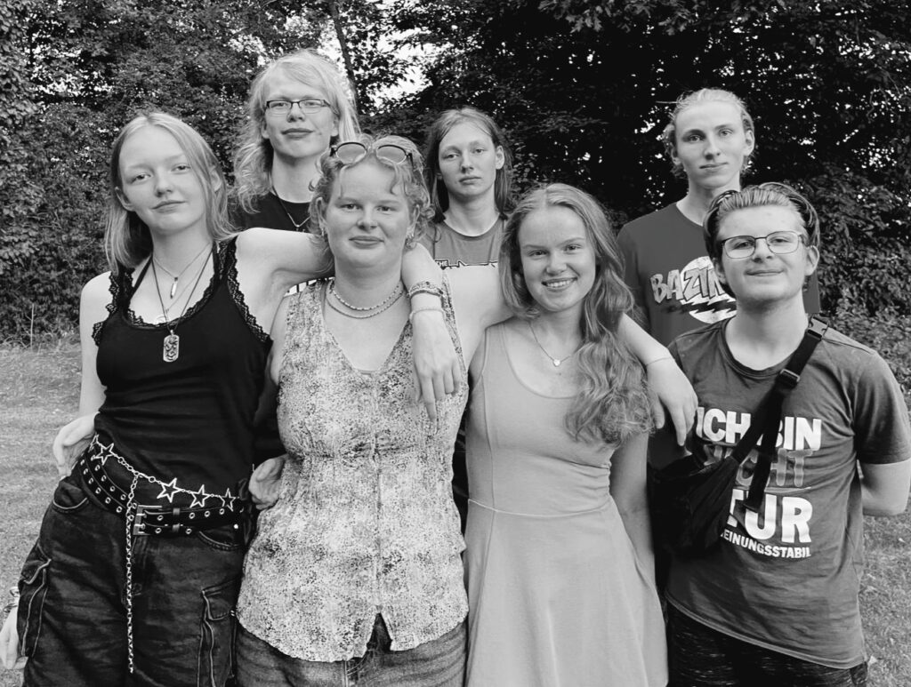 Gruppenfoto schwarz-weiß des aktuellen Ensembles AntiToxin, die Jugendlichen lächeln in die Kamera und haben den Arm um die jeweils andere Person gelegt