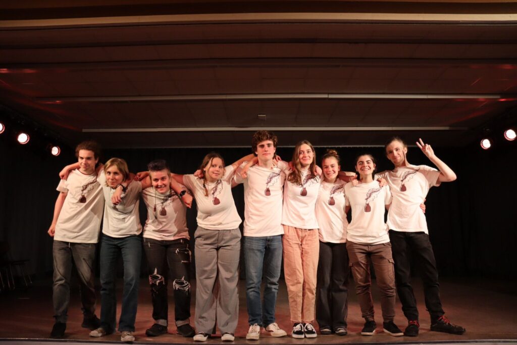 Das Ensemble von 2023 steht auf der Bühne in einer Reihe, die Arme um die Schultern des jeweils daneben stehenden gelegt, alle tragen weiße AntiToxin-T-Shirts