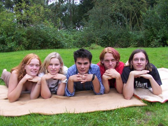 Gruppenfoto des Ensembles aus 2002, aufgenommen im Park, die fünf Jugendlichen liegen nebeneinander auf einer Decke und haben die Händer unterm Kinn verschränkt