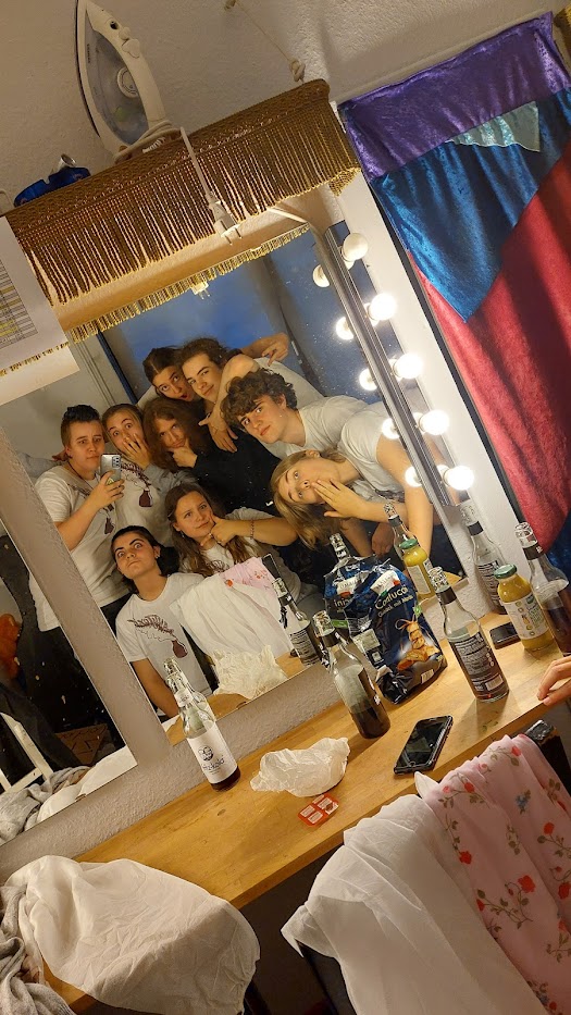 Eine Gruppe von neun jungen Menschen steht dicht beieinander vor einem beleuchteten Schminkspiegel und posiert fröhlich für ein Selfie. Sie tragen überwiegend weiße Oberteile, der Tisch vor ihnen ist mit Getränken, Chips und persönlichen Gegenständen bedeckt. Im Hintergrund sind bunte Vorhänge und ein aufgehängtes Bügeleisen zu sehen. Die Szene wirkt wie ein Blick in eine Künstlergarderobe oder ein Backstagebereich.