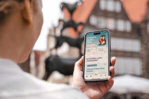 Ein Mensch hält ein Smartphone mit der neuen CityBuddy-App in der Hand im Hintergrund sind die Bremer Stadtmusikanten verschwommen zu erkennen