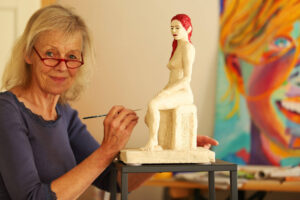 Das Foto zeigt die Bremer Künstlerin Birgit Heimburg, die lächelnd an einer Figur aus Pappmache arbeitet. 