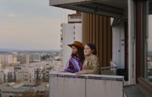 Zwei Mädchen stehen auf einem Balkon in einer Großstadt und blicken nachdenklich in die Ferne