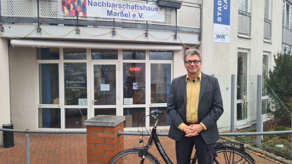 Quartiersmanager Marc Vobker steht vor seinem schwarzen Fahrrad am Nachbarschaftshaus in Marßel und lächelt in die Kamera.