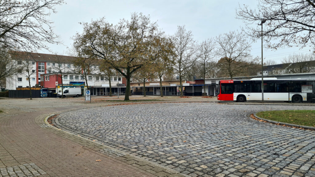 Helsingborger Platz in Marßel, Bus der BSAG rechts, der Platz ist leer, im Hintergrund steht ein weißer Van, die Ladenzeilen sind leer, nur das Geschäft ganz links hat geöffnet, ein türkischer Obsthändler.