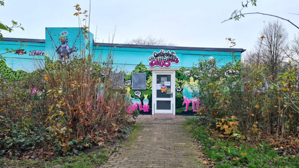 Frontale Aufnahme des Quartiertreffs "Bunte Mar", weiße Tür, die Wände sind bund bemalt, umrandet von Büschen