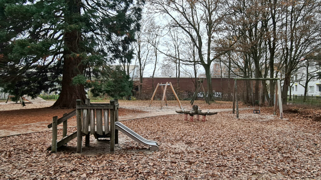 Ein ruhiger Spielplatz im Bremer Stadtteil Marßel nahe dem Marßeler Feld. Zu sehen sind eine kleine Holzrutsche, eine Wippe mit Federn, mehrere Schaukeln und große umstehende Bäume. Der Boden ist von Herbstlaub bedeckt, und im Hintergrund steht eine niedrige Mauer mit Graffiti.