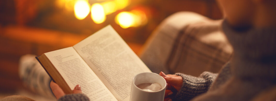 Jemand liest ein Buch mit einer Tasse in der Hand vor einem Kaminfeuer