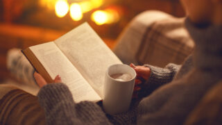 Jemand liest ein Buch mit einer Tasse in der Hand vor einem Kaminfeuer
