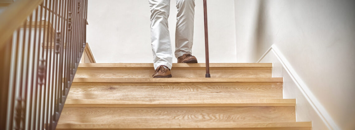 Älterer Herr geht eine Treppe mit einem Stock herunter, Nahaufnahme auf die Füße, Symbolbild für Pflegeimmobilien.