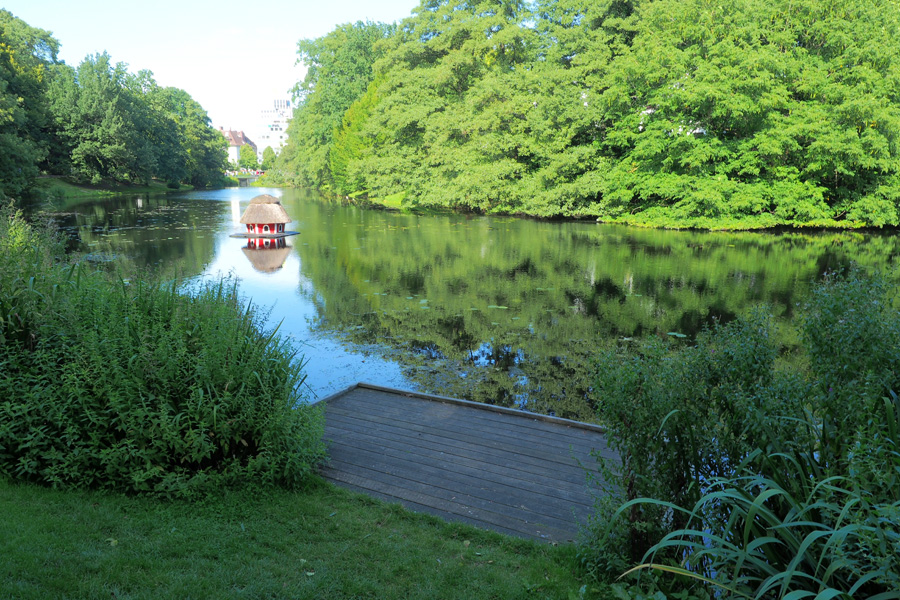 Holzsteg am Wasser in den Bremer Wallanlagen, umgeben von grüner Vegetation; auf dem Teich schwimmt ein kleines rotes Häuschen mit Reetdach.
