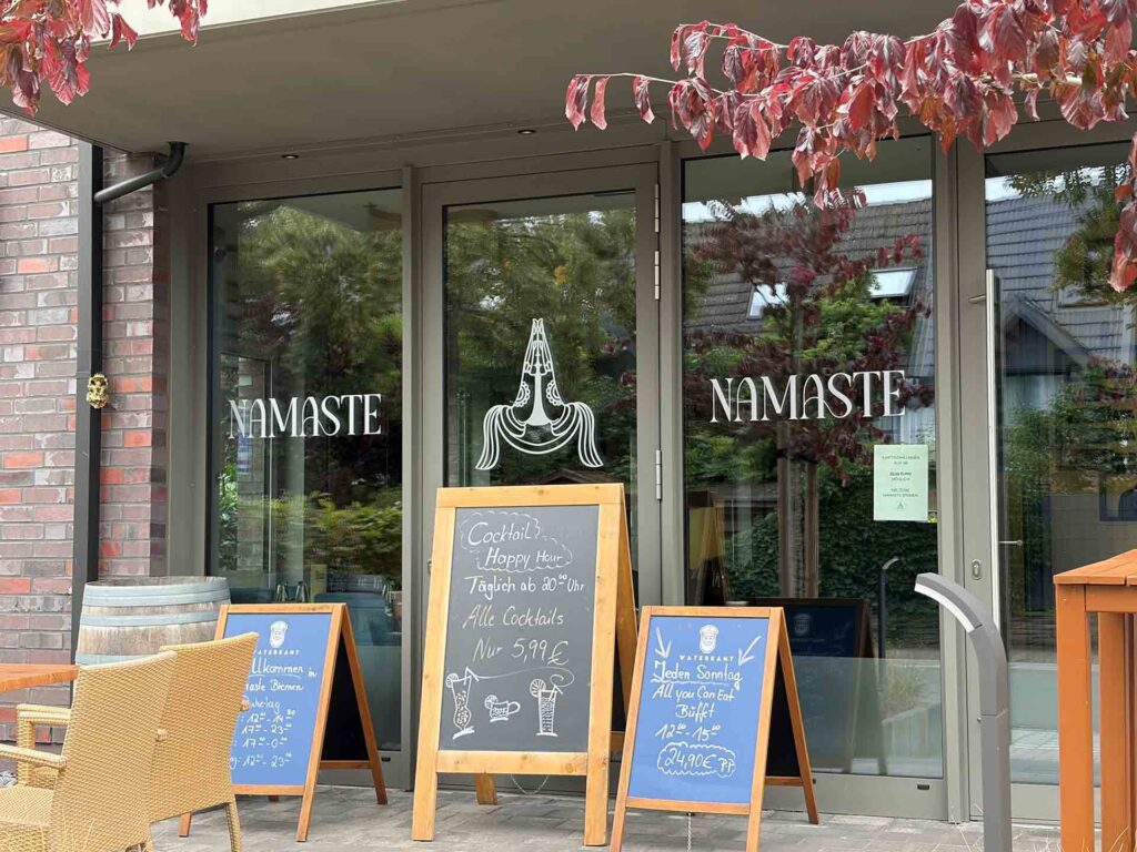 Gläserner Eingang des Restaurants Namaste mit Gebetshänden und mehreren Angebotstafeln